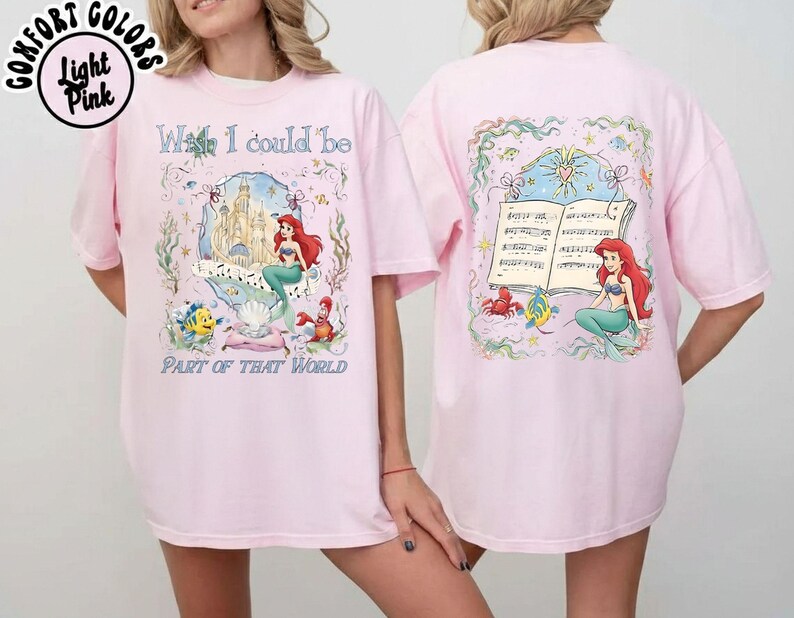 May include: Light pink t-shirt featuring a colorful graphic of a mermaid, castle, and text that reads "What I could be part of that world." The back of the shirt has a matching graphic of the mermaid and musical notes.