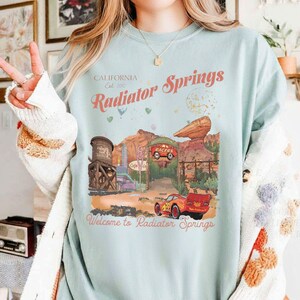 Puede incluir: Camiseta azul claro con un dise&ntilde;o de Radiator Springs, con texto que dice "California Est. 2012 Radiator Springs" y "Welcome to Radiator Springs". El dise&ntilde;o incluye un coche de dibujos animados y un paisaje des&eacute;rtico.