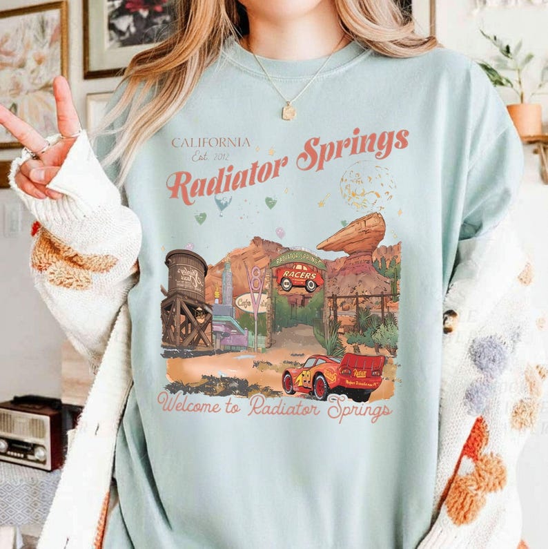 Puede incluir: Camiseta azul claro con un dise&ntilde;o de Radiator Springs, con texto que dice "California Est. 2012 Radiator Springs" y "Welcome to Radiator Springs". El dise&ntilde;o incluye un coche de dibujos animados y un paisaje des&eacute;rtico.