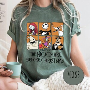 May include: Moss green t-shirt featuring a graphic of characters from "The Nightmare Before Christmas" in orange squares. The text "est.1993" and "THE NIGHTMARE BEFORE CHRISTMAS" are printed below the graphic.