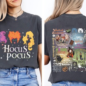 May include: Dark gray t-shirt featuring the text "Hocus Pocus" with illustrations of the Sanderson sisters' hair and a variety of themed graphics on the back. The shirt is a comfortable, casual style.
