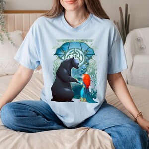 Op de afbeelding: Een lichtblauw T-shirt met een grafische print van een zwarte beer en een roodharig meisje. De beer steekt zijn poot uit naar het meisje, dat een blauwe jurk draagt. De print is omringd door een groen en blauw bloemenontwerp.