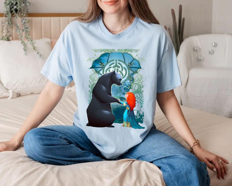 Op de afbeelding: Een lichtblauw T-shirt met een grafische print van een zwarte beer en een roodharig meisje. De beer steekt zijn poot uit naar het meisje, dat een blauwe jurk draagt. De print is omringd door een groen en blauw bloemenontwerp.