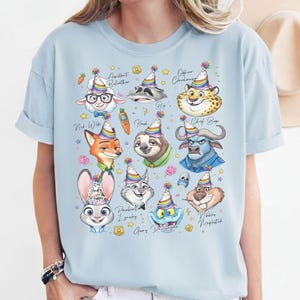 Puede incluir: Camiseta azul claro con personajes de dibujos animados de Zootopia, cada uno con un gorro de fiesta. Los personajes incluyen a Judy Hopps, Nick Wilde y Flash. La camiseta tiene el texto "Judy Hopps" y otros nombres de personajes.