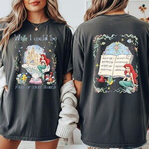 May include: Dark gray t-shirt featuring a colorful illustration of Ariel from The Little Mermaid. The front of the shirt has the text "Wish I could be Part of That World" with a castle and sea creatures. The back has Ariel with sheet music.