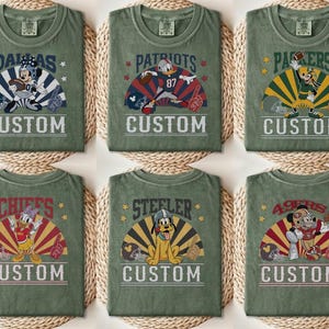 Op de afbeelding: Zes olijfgroene t-shirts met verschillende teamnamen en stripfiguren. Elk shirt heeft een teamnaam, een stripfiguur in een voetbaluniform en het woord "CUSTOM" in witte letters. De shirts zijn gerangschikt op een geweven oppervlak.