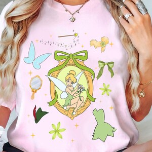 Op de afbeelding: Roze T-shirt met een groen en gouden frame met Tinkerbell die een bloem vasthoudt. Het frame is omgeven door groene strikken, sterren en andere sprookjesachtige afbeeldingen.
