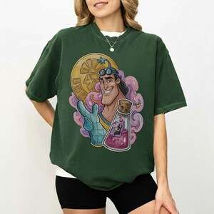 May include: Dark green t-shirt featuring a cartoon character with goggles, holding a gold coin and a potion bottle. The graphic includes pink and purple swirls. The shirt is a relaxed fit.
