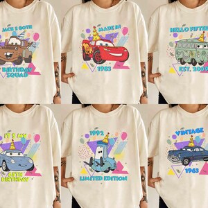 May include: Six white t-shirts with different cartoon car designs and text. The designs include a red car with the text "Made in 1983", a blue car with the text "1992 Limited Edition", a green van with the text "Hello Fifteen Est. 2009", a blue car with the text "Vintage 1963", a blue car with the text "It's My 45th Birthday", and a tow truck with the text "Jack's Goth Birthday Squad".