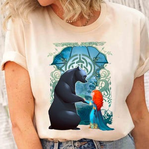 Op de afbeelding: Een wit T-shirt met een grafische print van een beer en een meisje met rood haar. De beer is zwart en het meisje draagt een blauwe jurk. De print is tegen een blauwe en groene achtergrond met een draaiende print.