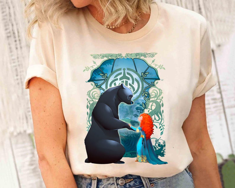 Op de afbeelding: Een wit T-shirt met een grafische print van een beer en een meisje met rood haar. De beer is zwart en het meisje draagt een blauwe jurk. De print is tegen een blauwe en groene achtergrond met een draaiende print.