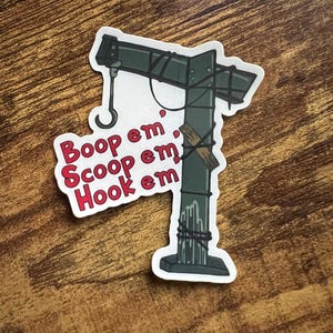 May include: A sticker featuring a stylized crane design in shades of green and black, with a curved hook. The sticker includes the text "Boop em, Scoop em, Hook em" in red letters.
