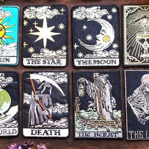 May include: A set of eight rectangular embroidered patches featuring tarot card designs. The patches depict The Sun, The Star, The Moon, The Ace of Cups, The World, Death, The Hermit, and The Lovers, each with detailed black, white, and gold thread work.