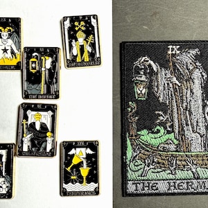 May include: A collection of six rectangular enamel pins featuring tarot card designs including The Devil, The Hermit, The Hierophant, The Emperor, and Temperance, alongside one large rectangular embroidered patch depicting The Hermit tarot card.