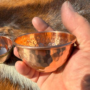May include: A small, hammered copper bowl with a rounded shape. The bowl has a textured surface with a series of small, circular indentations. The copper has a warm, reddish-brown color. The bowl is held in a hand.