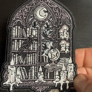 May include: An embroidered patch in a gothic arch shape featuring a library scene with bookshelves, a raven, a globe, and multiple lit candles. The design is rendered in shades of black, gray, and white with a crescent moon at the top.