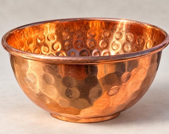 May include: A round, metallic copper bowl featuring a distinct hammered texture across its entire exterior and interior surface. The bowl has a smooth, rounded rim and a polished finish that reflects light, showcasing the warm reddish-brown color.
