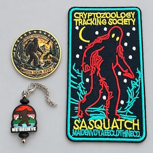 May include: A circular metal coin featuring a Sasquatch, a rectangular embroidered patch with a red Sasquatch silhouette and the text Cryptozoology Tracking Society, and a small rubber charm with a Sasquatch graphic and the text We Believe.