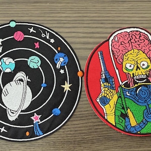 May include: Two embroidered patches. One is a black circle with a white outline of a solar system, planets, and stars. The other is a red circle with a yellow-skinned alien holding a ray gun. The alien has a large brain and is wearing a green and blue uniform.