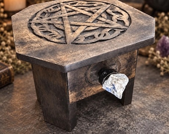 May include: A small, dark brown wooden box with a hexagonal top featuring a carved pentagram design. The box stands on four short legs and includes a single drawer front adorned with a clear, rough-cut crystal knob that serves as a handle.