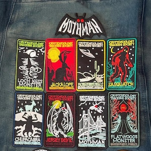 May include: A collection of nine embroidered patches on denim fabric. One bat-shaped patch reads MOTHMAN. Eight rectangular patches feature cryptozoology themes including Loch Ness Monster, Jackalope, Extraterrestrials, Sasquatch, Chupacabra, Jersey Devil, Mothman, and Flatwoods Monster.
