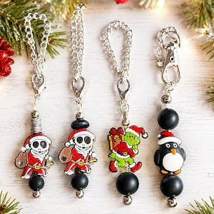 May include: Four silver-toned metal keychains featuring enamel charms of holiday characters including two skeletons in Santa suits, a green creature carrying a gift, and a penguin wearing a Santa hat, accented with black beads and silver chains.