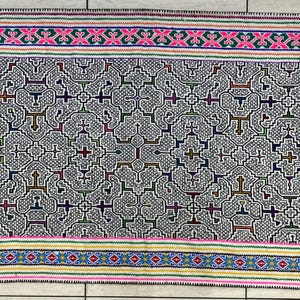 May include: A white woven tapestry with a geometric pattern in shades of blue, green, brown, pink, and yellow. The pattern is made up of repeating crosses and squares.