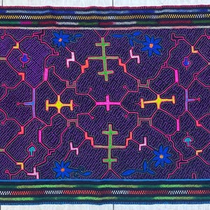 May include: A rectangular textile with a dark purple background, featuring a repeating pattern of crosses and floral elements in various colors. The edges are bordered with colorful striped patterns.