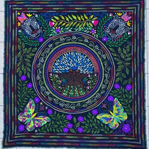 May include: A square, dark blue blanket with a vibrant embroidered design. The design features a central circular motif with a peacock-like figure, surrounded by butterflies, floral elements, and stylized jaguar faces. The blanket has a detailed, colorful border.