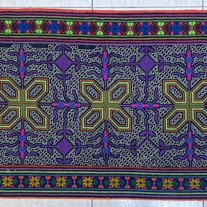 May include: A rectangular textile with a repeating geometric pattern. The design features large, four-leaf clover-like shapes in green and yellow, surrounded by purple and blue details. The borders have floral and geometric patterns in various colors.