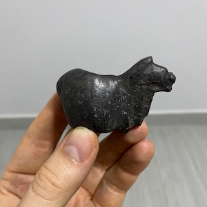 Puede incluir: Una pequeña escultura gris oscura de un cerdo. El cerdo está de pie, con la cabeza ligeramente levantada. La escultura tiene una superficie rugosa y texturizada y está sostenida en una mano.