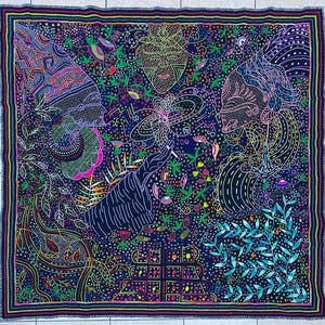 May include: A square, dark blue textile with a colorful, intricate design. The artwork features stylized faces, hands, and botanical elements in shades of pink, green, yellow, and purple. The border is a multi-colored zig-zag pattern.