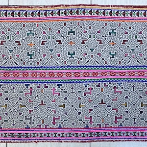 May include: A rectangular textile with a repeating geometric pattern in gray, green, red, and blue. The borders are trimmed with pink and brown. The textile is likely hand-woven.