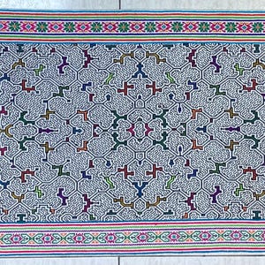 May include: A rectangular textile with a repeating geometric pattern in white, red, blue, green, and yellow. The borders feature bands of color and floral designs. This decorative textile could be used as a table runner or wall hanging.