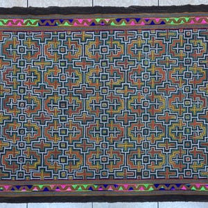 May include: Rectangular textile with a geometric pattern in shades of brown, orange, yellow, and blue. The design features cross-like shapes. The border is dark brown with a colorful trim of pink, green, and blue.