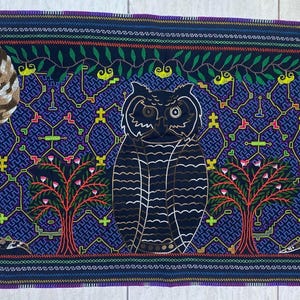 May include: A rectangular textile with a dark blue background featuring an owl, two serpent-like creatures, and trees. The owl is black and white, the creatures have brown and white wings, and the trees have red and green leaves. The border is a mix of colors.