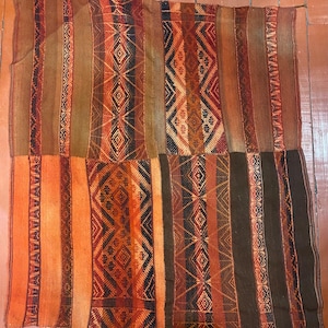 Könnte beinhalten: Ein gewebtes Textil mit einem geometrischen Muster in Orange-, Braun- und Schwarztönen. Das Textil weist vertikale Streifen und Rauten auf, wodurch ein strukturiertes und farbenfrohes Design entsteht. Der obere Teil ist ärmellos.