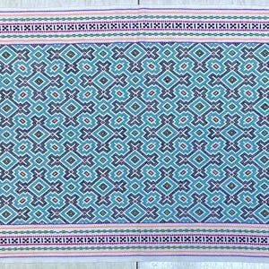 May include: A rectangular textile with a repeating geometric pattern in shades of teal, purple, and brown. The design is framed by borders of pink, green, and purple stripes, and a white background. The textile appears to be hand-woven.
