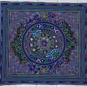 May include: A square, intricately designed blanket with a dark blue background. The blanket features a central circular pattern with floral and geometric elements in shades of purple, green, and pink. Butterflies and leaves are also incorporated into the design.
