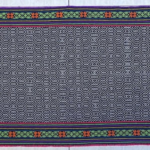 May include: A rectangular, woven textile with a dark blue background and a repeating geometric pattern. The edges feature bands of color, including green, orange, red, and purple, creating a decorative border. The textile is likely used as a table runner or decorative cloth.