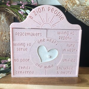 May include: A pink decorative plaque with the words "A Zion People" at the top. The plaque has a heart-shaped cutout in the center and includes phrases like "Peacemakers" and "Pure in Heart."