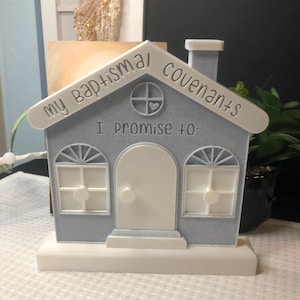 Pode incluir: Um objeto decorativo em forma de casa azul clara com o texto "my baptismal covenants" e "I promise to:". A casa tem uma porta, janelas e acabamentos brancos. Uma pequena chaminé é visível no telhado. O objeto está sobre uma base branca.