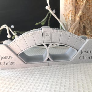 May include: A gray decorative bridge with the words "Jesus Christ" on each side. The bridge has an arched design with a small rectangular plaque in the center. The bridge is made of a stone-like material.