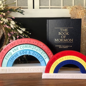 May include: Two rainbow-shaped decorative objects with white bases labeled 'my baptismal covenant' and 'my baptismal covenants'. The rainbows feature engraved text including 'Give me the Gift of the Holy Ghost', 'keep the commandments', and 'serve God & Others'. A black book titled 'THE BOOK OF MORMON' stands behind them.