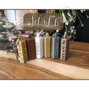 May include: A set of six rectangular keychains in various colors, including gold, brown, white, and gray. Each keychain has text in different languages. The keychains are displayed on a wooden surface.