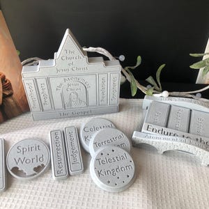 May include: A collection of gray religious-themed decorative pieces. Includes a building-shaped piece with text "The Church of Jesus Christ", and other pieces with words like "Spirit World", "Resurrection", and "Endure to the End".