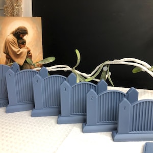Puede incluir: Una colección de seis decoraciones de lápidas en miniatura de color azul claro. Cada lápida tiene un diseño único, con los números del 1 al 5 visibles. Una pintura de Jesús y un niño está en el fondo, junto con vegetación decorativa.