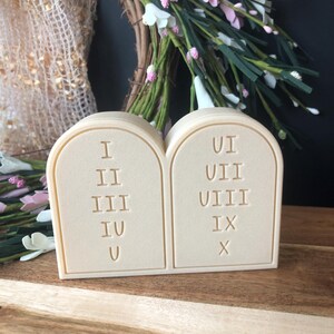 May include: A beige-colored Ten Commandments decorative piece. The two tablets are side-by-side, with Roman numerals I through X. The background includes a floral wreath with pink and white flowers.