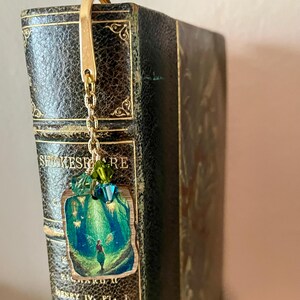 Könnte beinhalten: Ein goldfarbener Lesezeichen mit einer Kette und einem kleinen, rechteckigen Anhänger mit einer grün-blauen Waldszene. Das Lesezeichen ist an einem alten Buch mit dem Titel "SHAKESPEARE" befestigt.