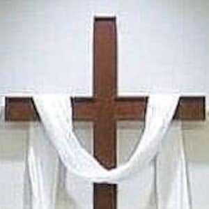 May include: A wooden cross with a white cloth draped over it. The cross is mounted on a white wall.